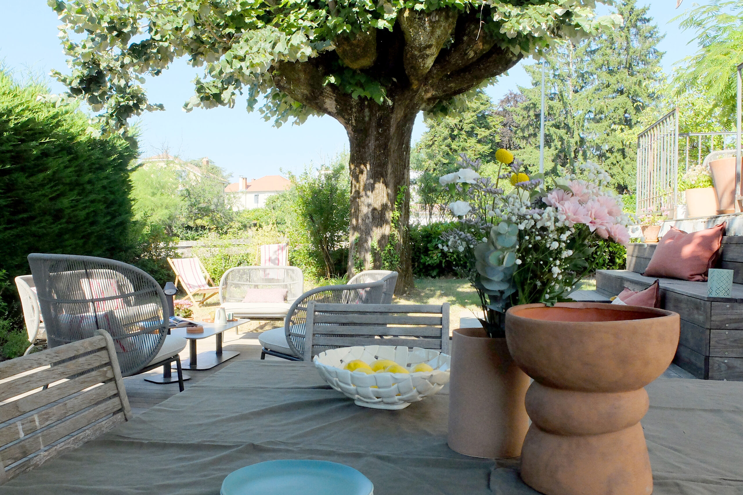 Terrasse en bois et salon de jardin sous un arbre, bordés de plantations fleuries.